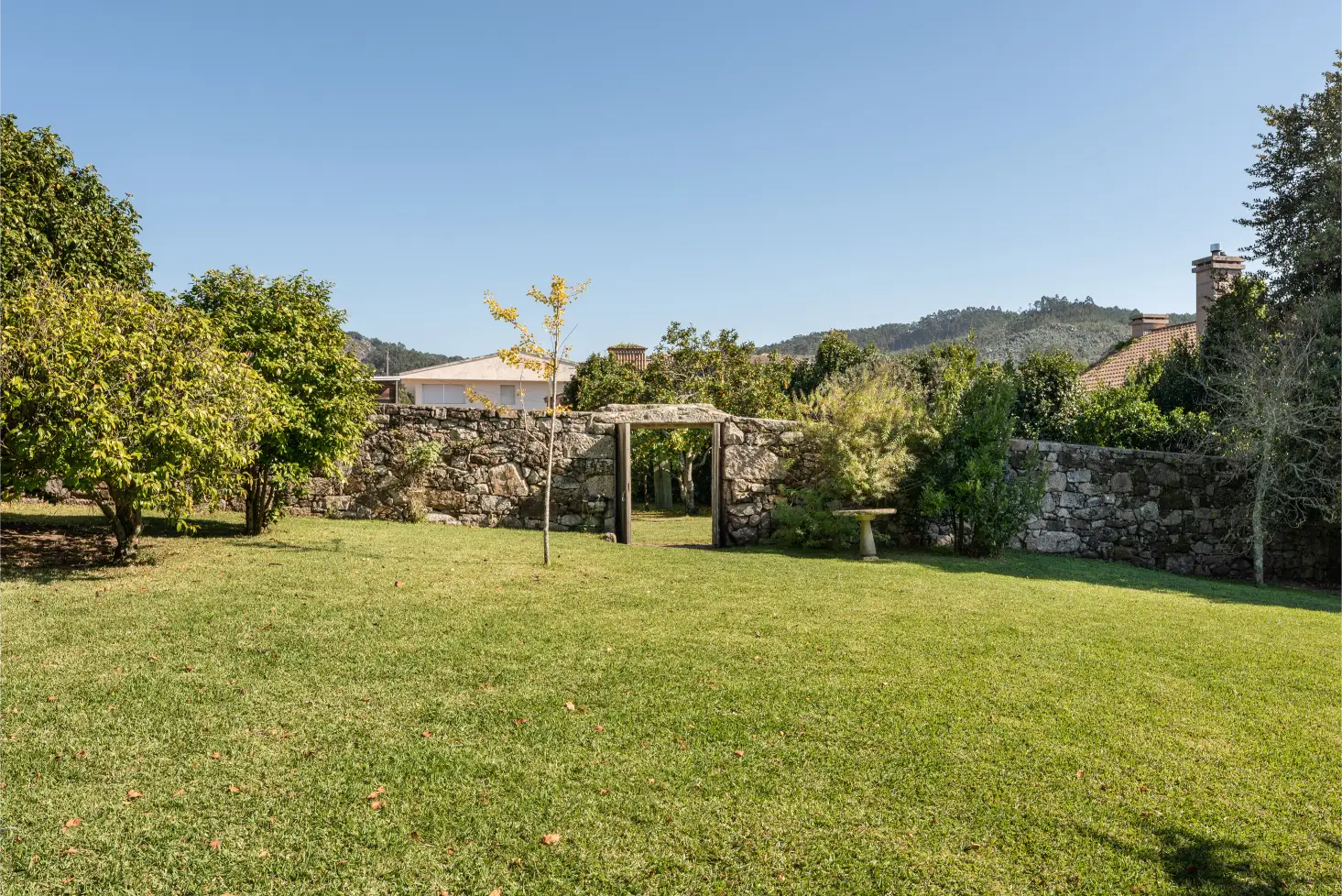 Stone wall with an open wooden gate surrounded by green trees and grass under a clear blue sky.