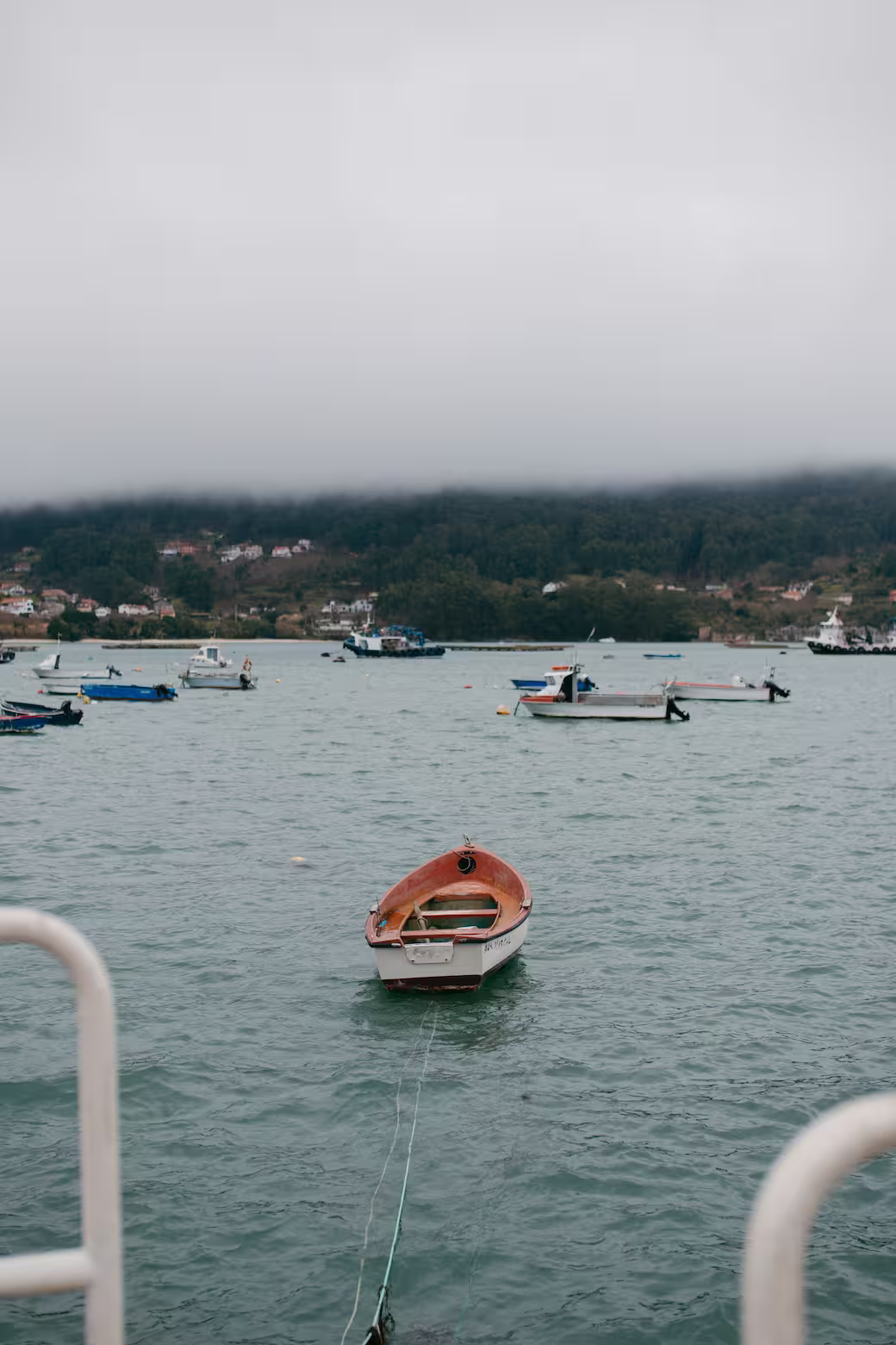 Small red and white boat tied to a dock on calm water with other boats and a fog-covered hillside in the background.