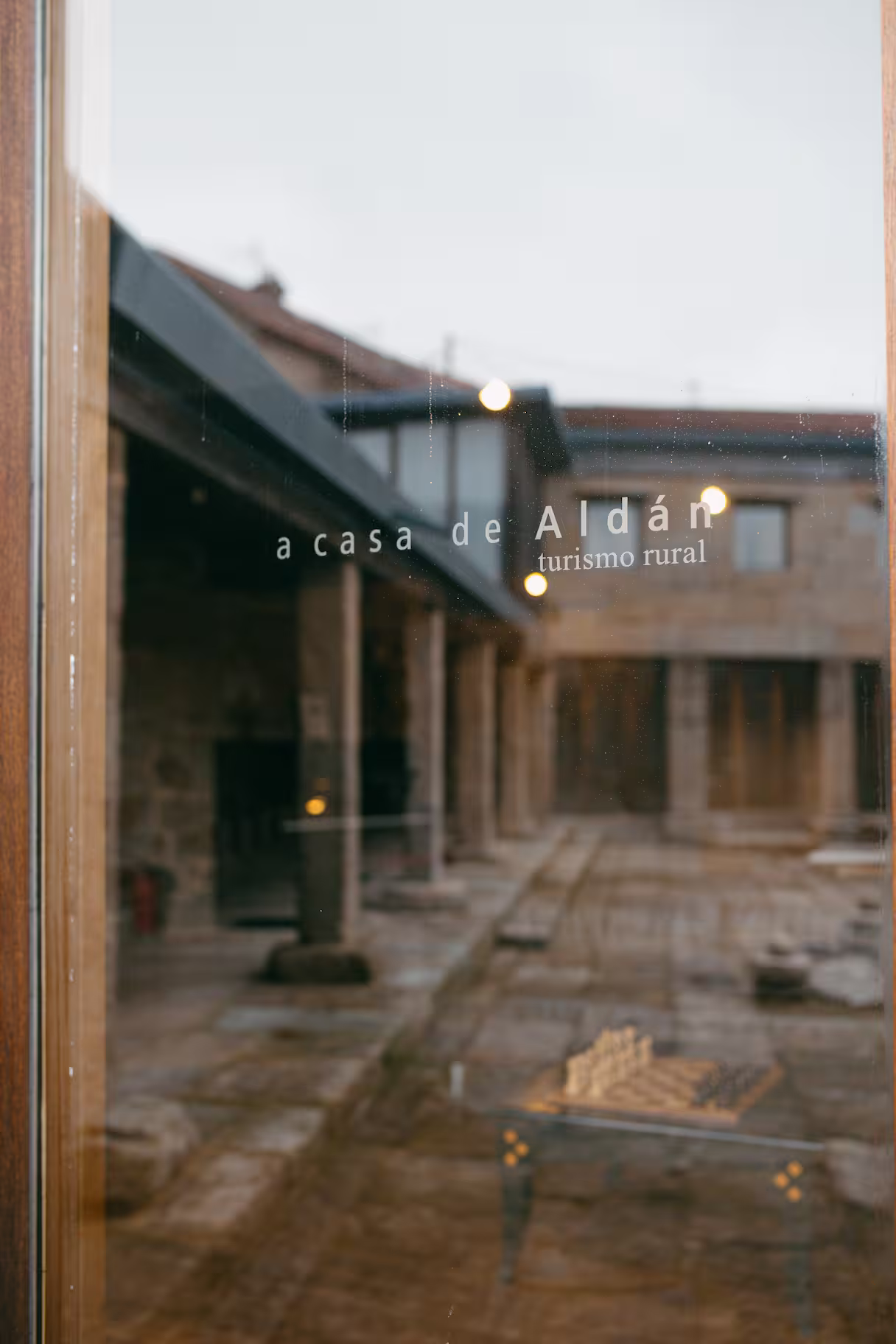 View through glass door showing an outdoor courtyard with stone columns and tiled floor, with text 'a casa de Aldán turismo rural' on the glass.