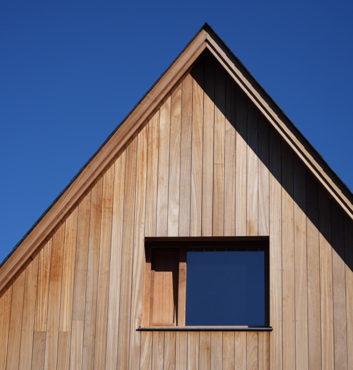 Façade en bois clair d'une maison avec un toit en pente et une fenêtre carrée sous un ciel bleu.