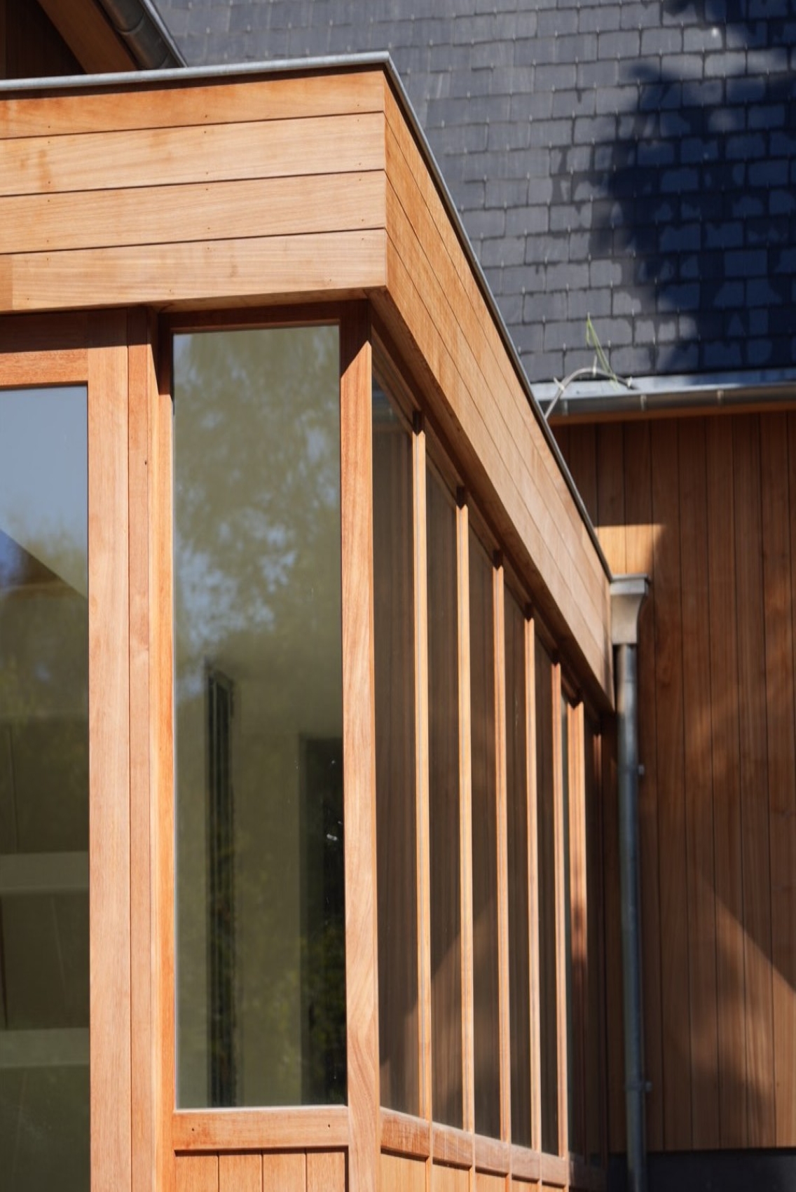 Angle d'une maison moderne avec bardage en bois et grandes fenêtres vitrées sous un toit en ardoise.