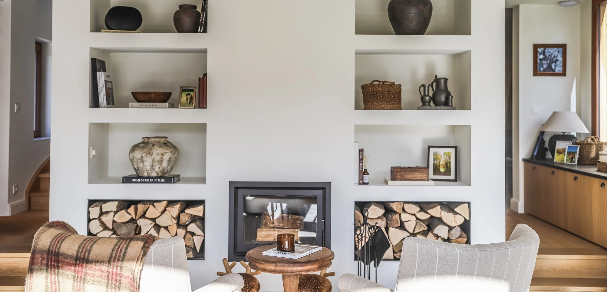 Salon lumineux avec cheminée encadrée de niches intégrées contenant des livres, vases, cadres photos, et bûches de bois, avec deux fauteuils et une table basse en bois.