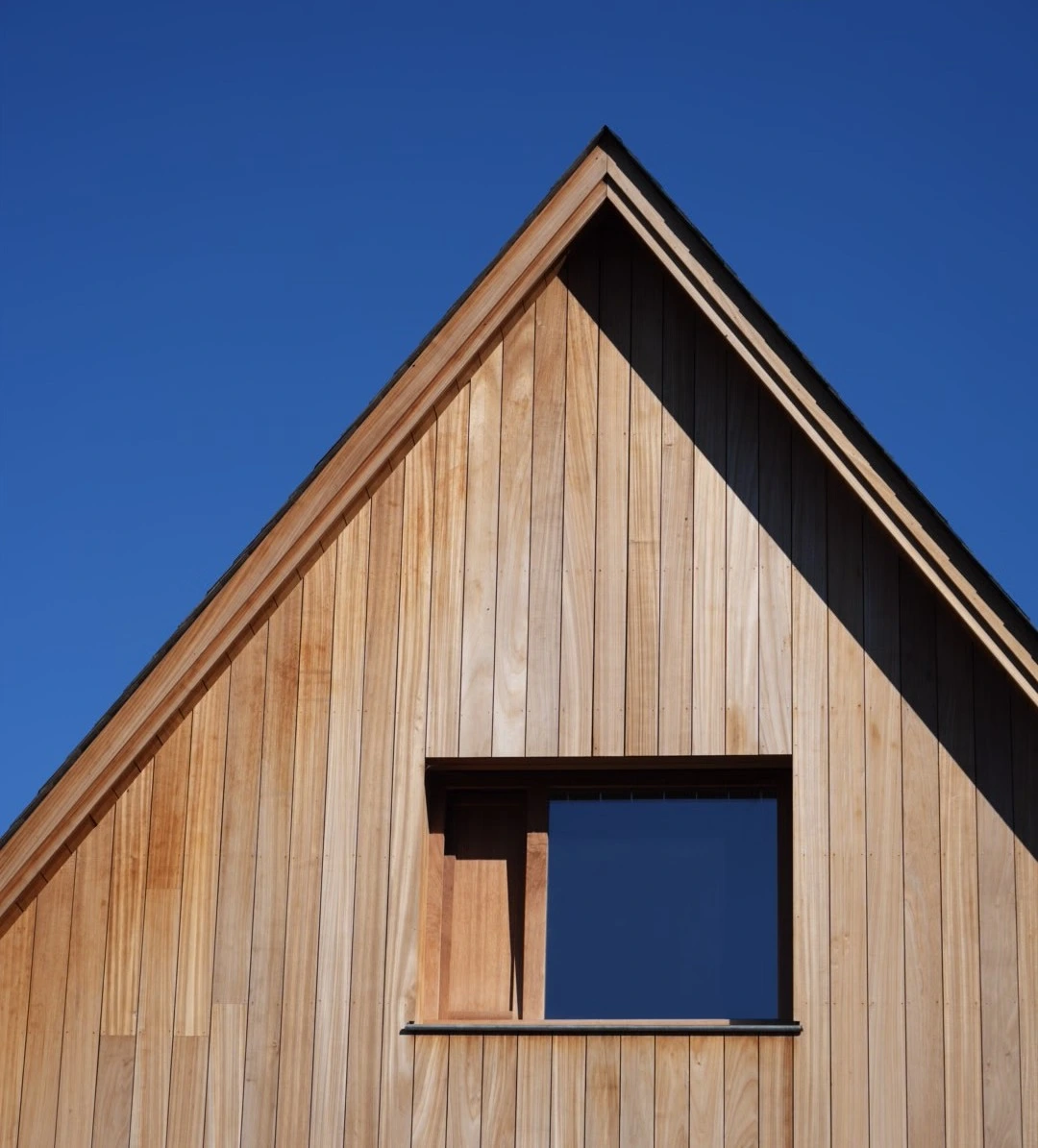 Façade en bois clair d'une maison avec un toit en pente et une fenêtre carrée sous un ciel bleu.