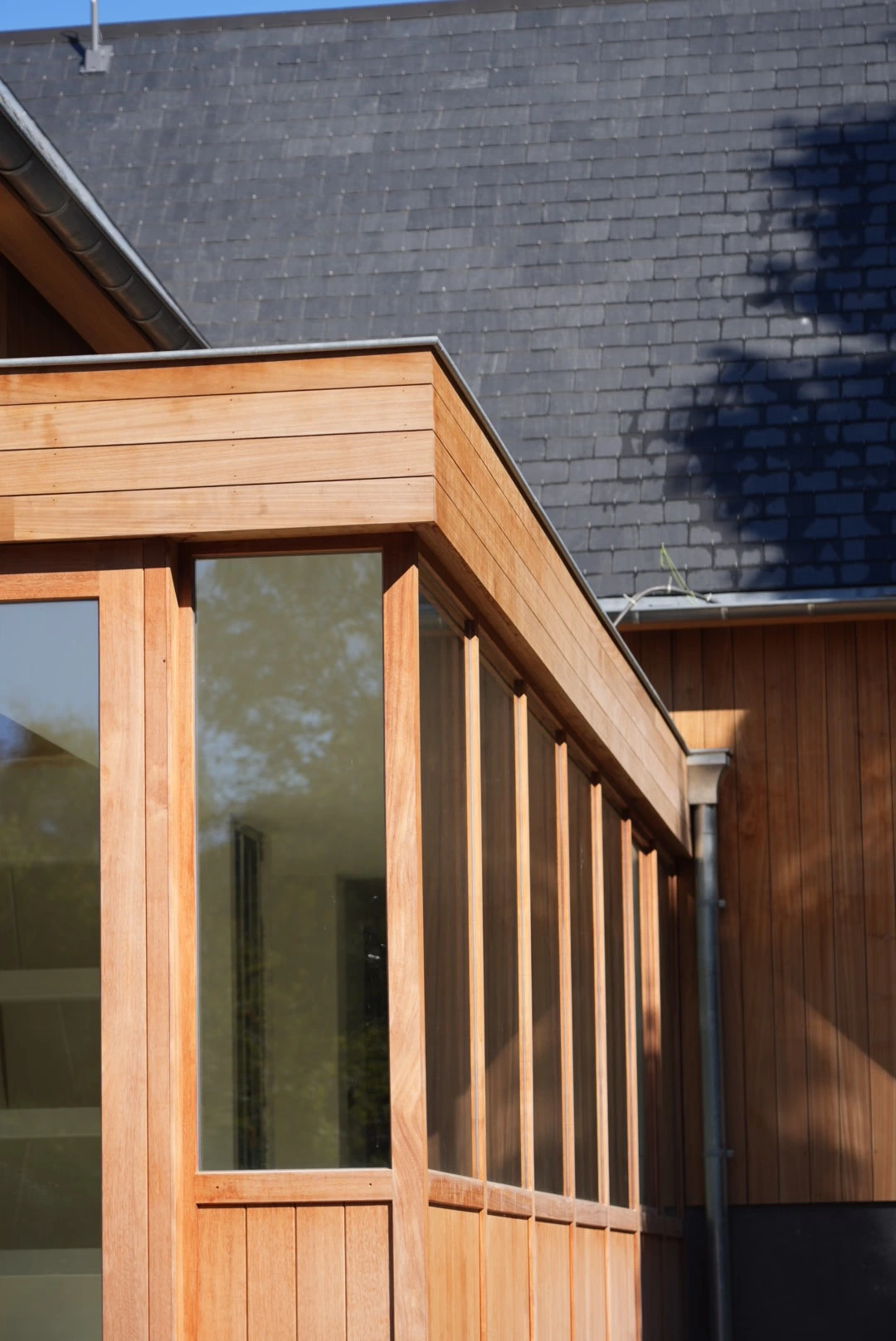 Angle d'une maison moderne avec bardage en bois et grandes fenêtres vitrées sous un toit en ardoise.