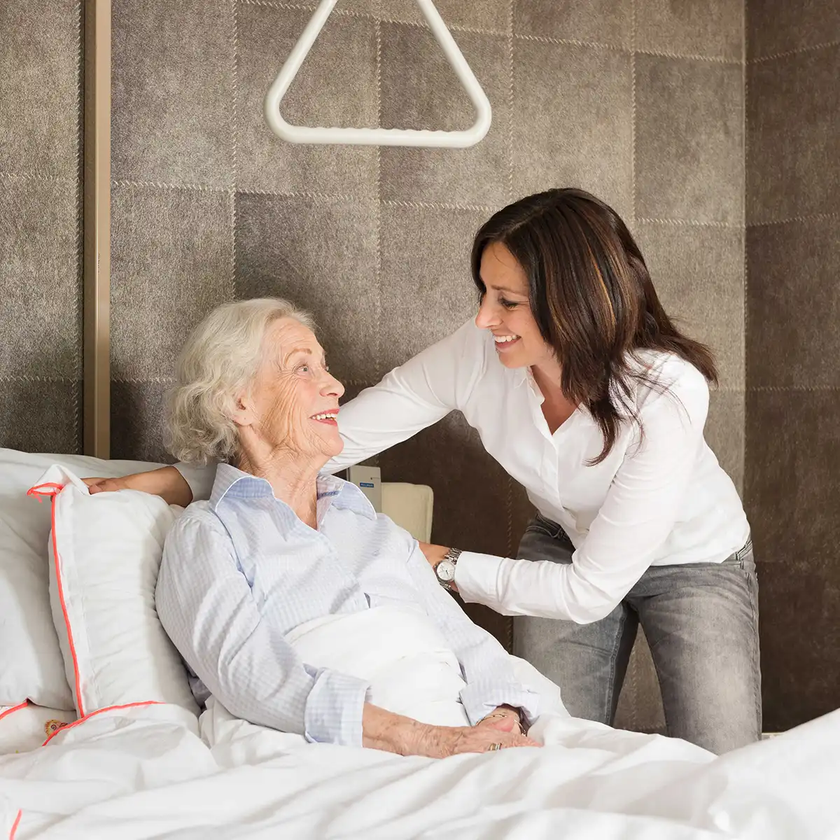 Een oudere vrouw zit in bed en glimlacht naar een jongere vrouw die haar bemoedigend vasthoudt.