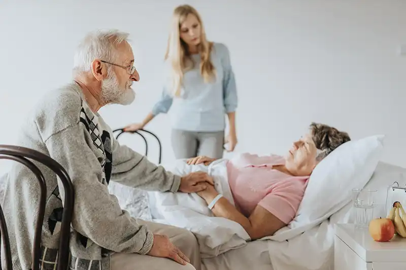 Oudere man die hand van zieke vrouw houdt die in bed ligt, terwijl een jonge vrouw bezorgd kijkt.