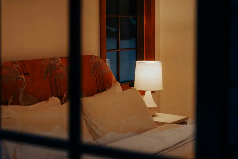 Slaapkamer met bed dat kussens en een lamp op een nachtkastje heeft, gezien door een raam.