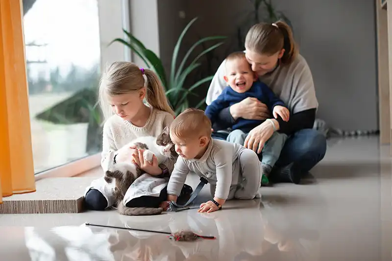 Vrouw zit op de vloer met twee jonge kinderen, terwijl een meisje een kat vasthoudt en een baby op de vloer speelt.