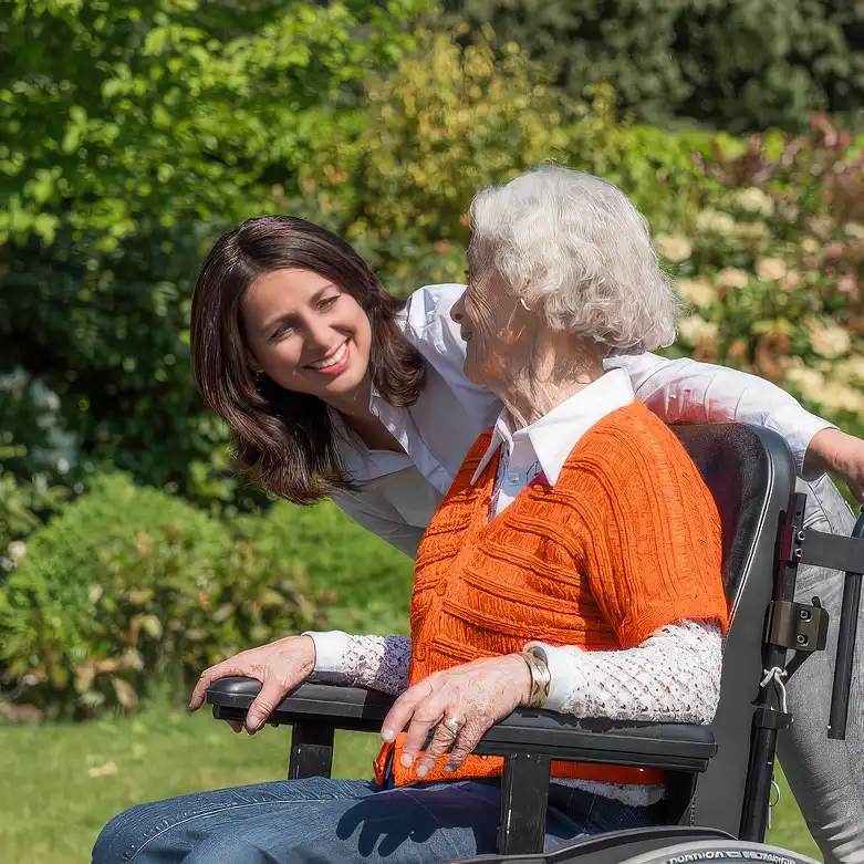 Jonge vrouw lacht en praat met oudere vrouw in rolstoel in een zonnige tuin.