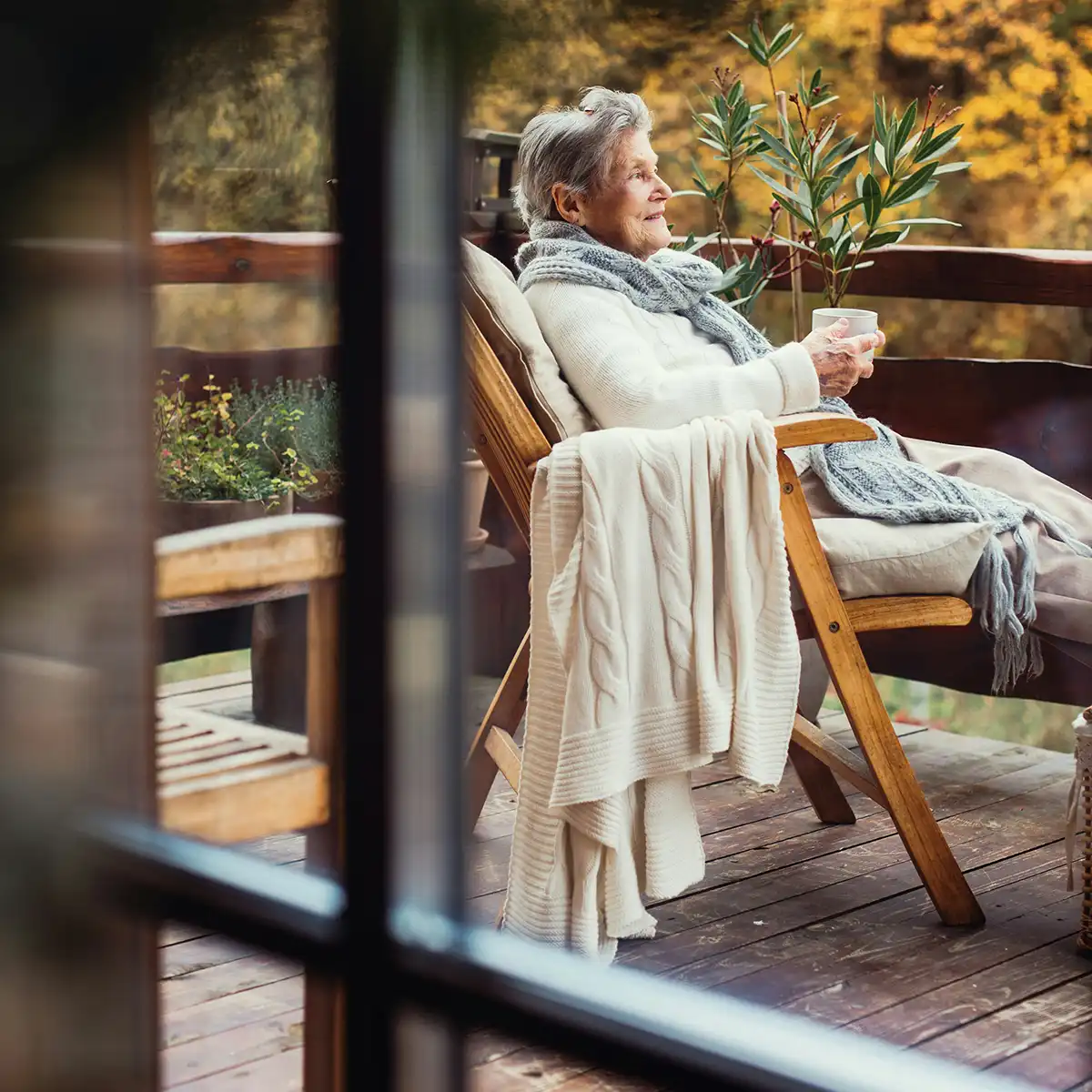 Oudere vrouw ontspannen in een leren stoel op een houten terras met een kopje thee, omringd door planten.