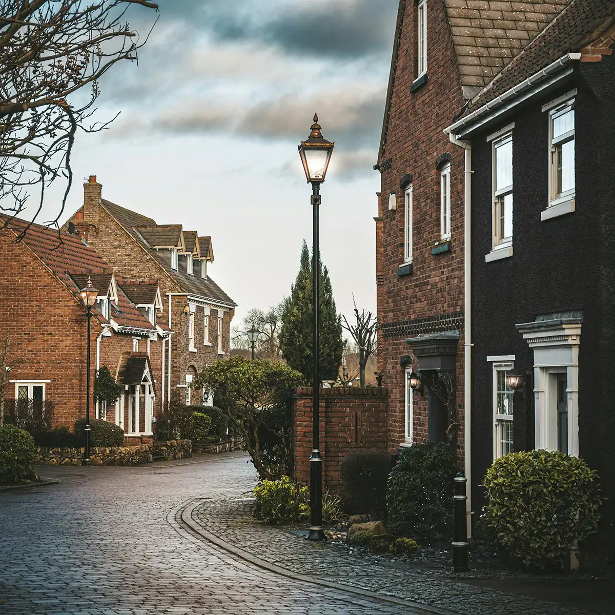 Verlichte straatlantaarns langs een kronkelende straat met bakstenen huizen en struiken bij schemering.