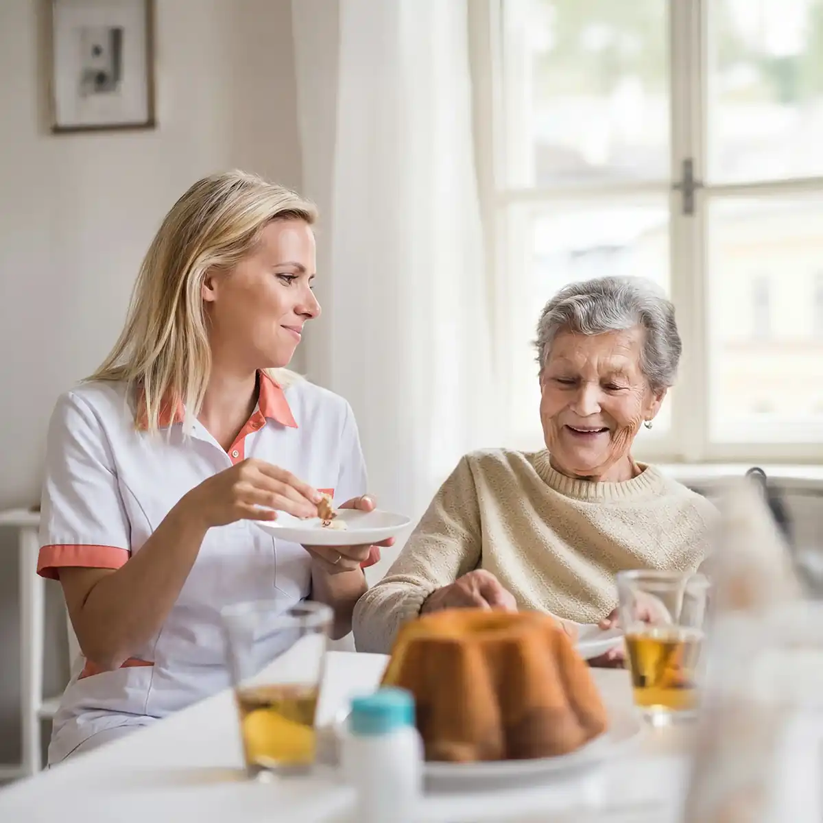 Verzorgster die een oudere vrouw helpt eten aan een tafel met drinken en cake in een lichte kamer.