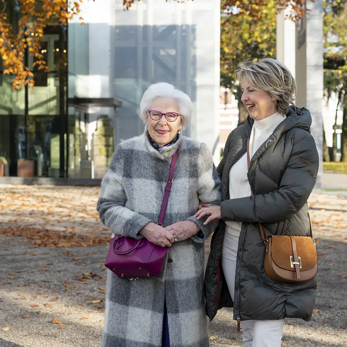Twee vrouwen, een oudere en een jongere, wandelen buiten in herfstachtige omgeving terwijl ze lachen en elkaar vasthouden.