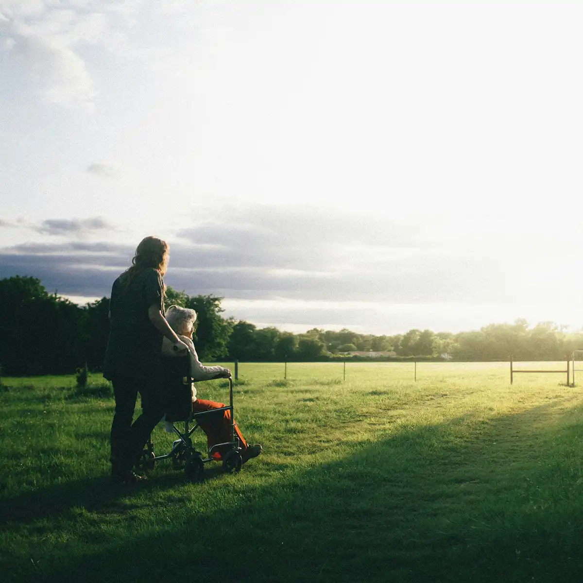 Verzorger duwt oudere vrouw in rolstoel over grasveld bij zonsondergang.