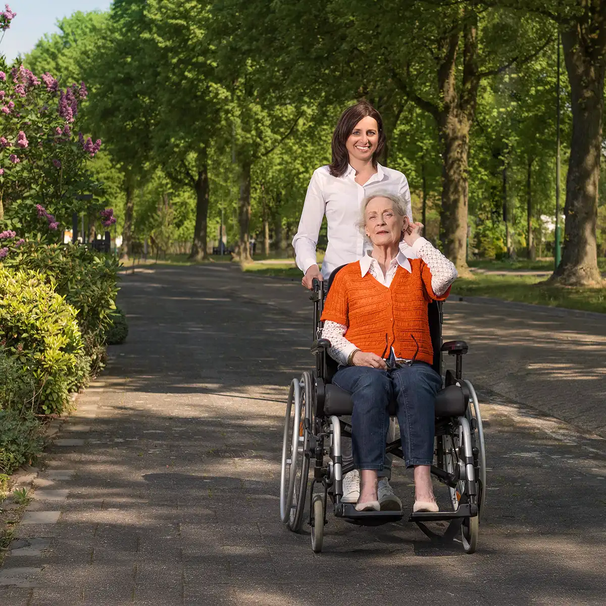 Een oudere vrouw in een rolstoel wordt buiten door een jonge vrouw met witte blouse duwende op een pad omgeven door bomen en struiken.
