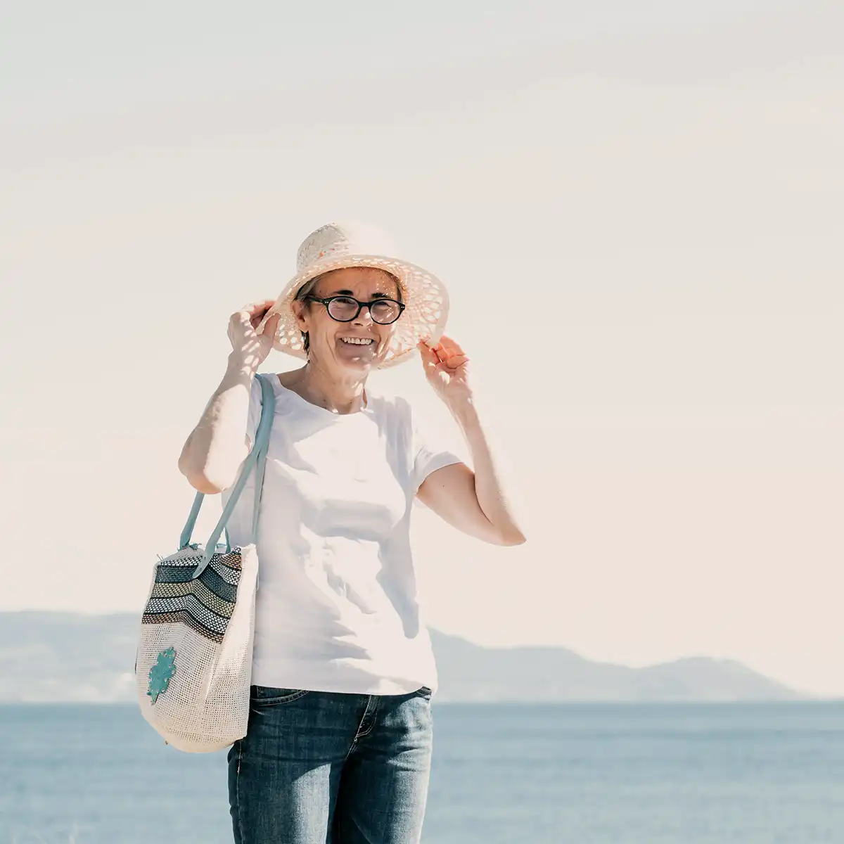 Vrouw met zonnehoed en zonnebril staat lachend bij zee met een tas over haar schouder.