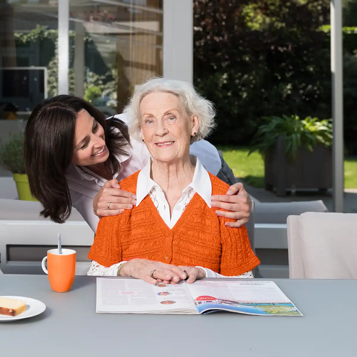 Een oudere vrouw in een oranje trui zit aan een tafel met een open boek, terwijl een jongere vrouw haar vriendelijk omarmt van achteren.