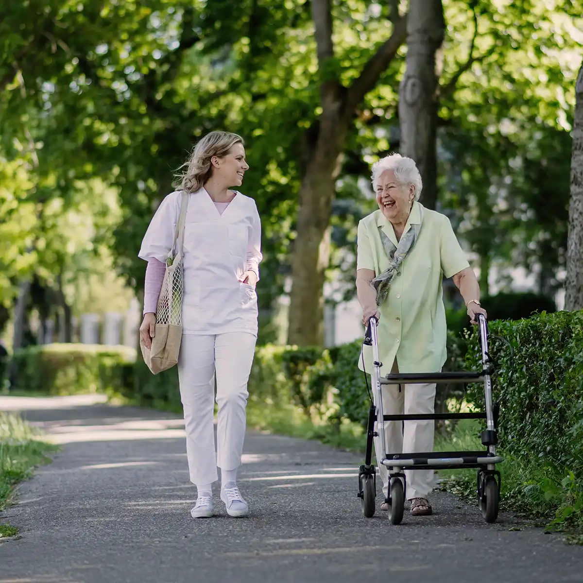 Een zorgverlener loopt buiten op een pad samen met een lachende oudere vrouw die een rollator gebruikt.