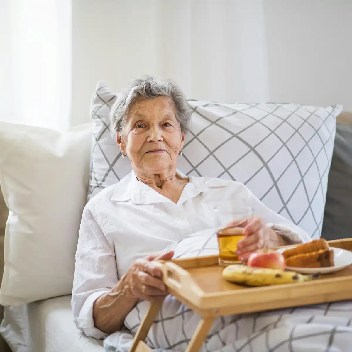 Oudere vrouw zit in bed met een dienblad met fruit, een glas sap en een bord met snacks.