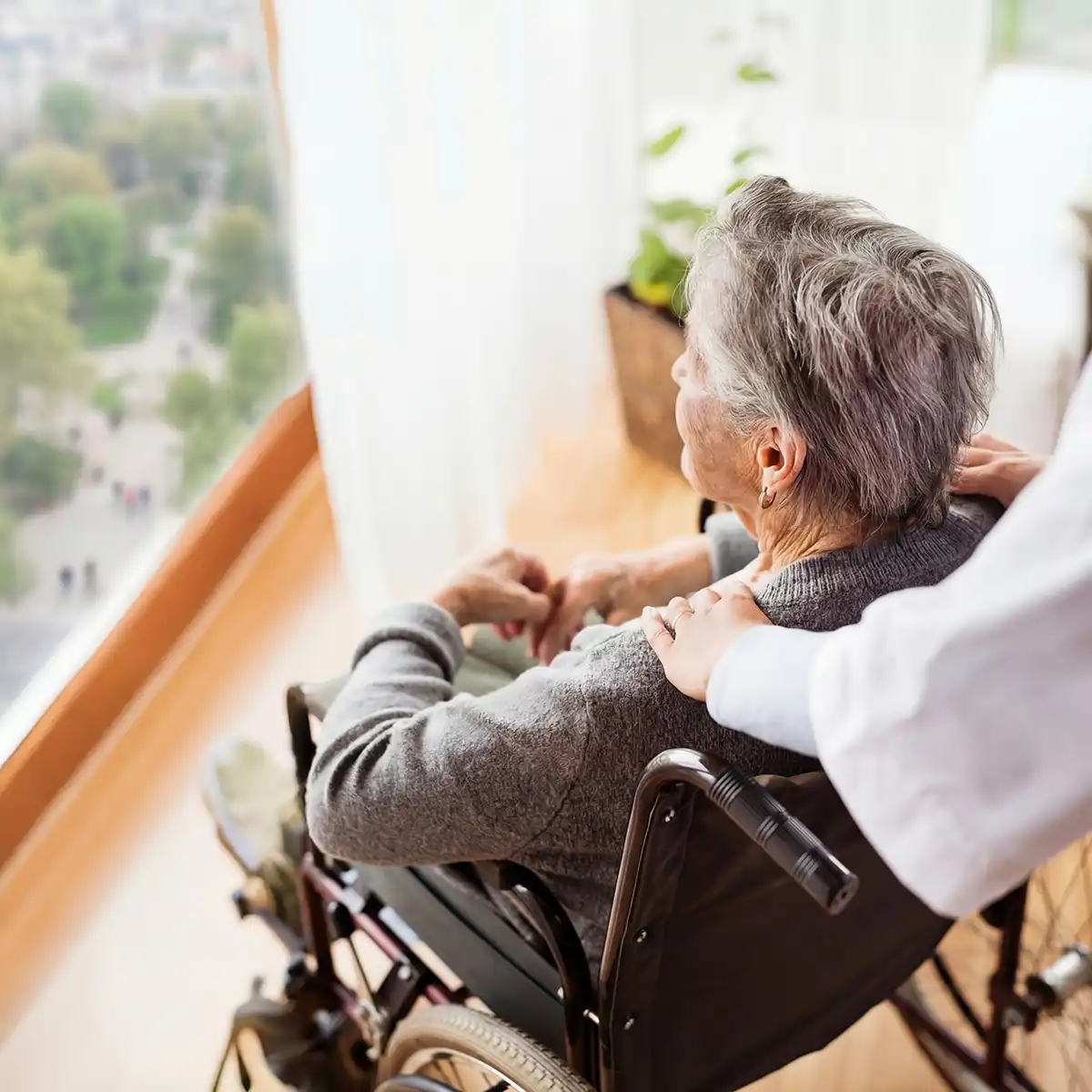 Een oudere vrouw in een rolstoel kijkt naar buiten terwijl een verzorger haar vriendelijk op de schouder raakt.