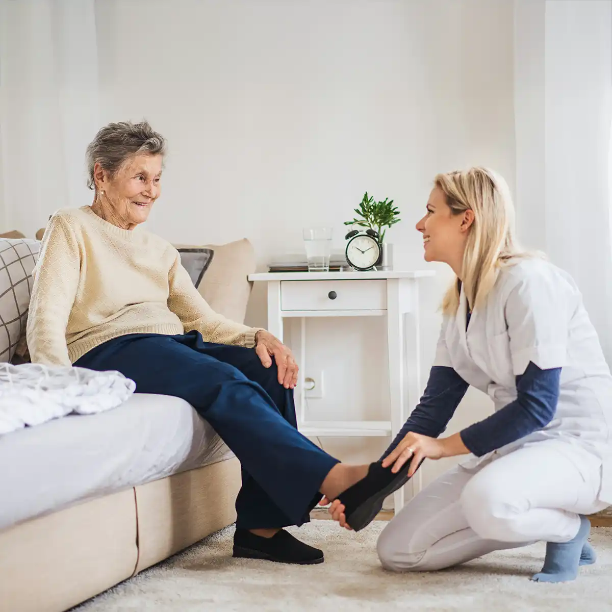 Verpleegkundige helpt oudere vrouw met het aandoen van haar schoen terwijl ze glimlachen in een huiselijke omgeving.