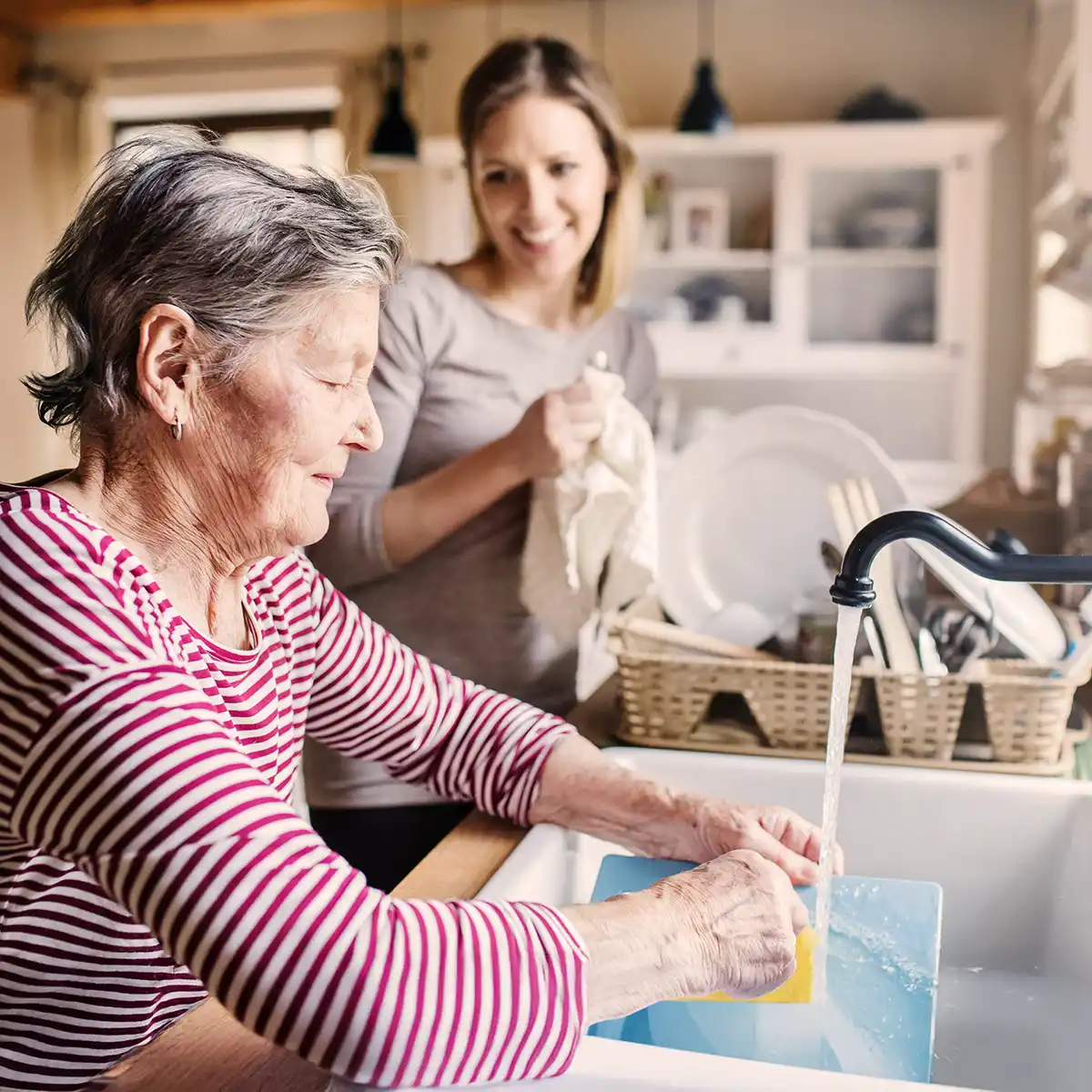 Oudere vrouw wast een snijplank in de gootsteen terwijl een jongere vrouw met een doek glimlacht in een keuken.