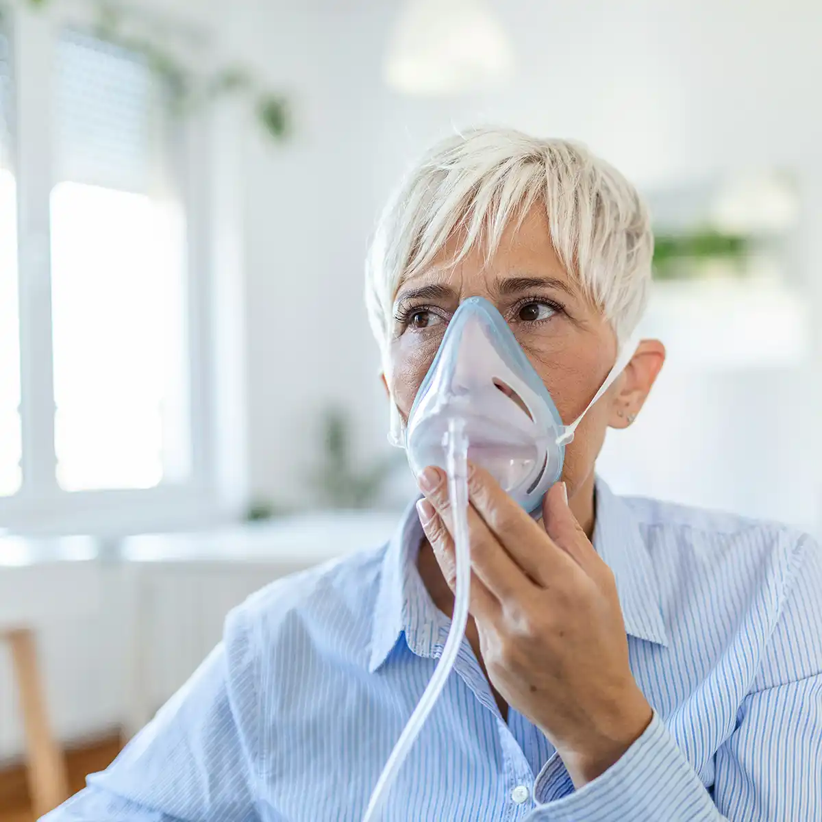 Een oudere vrouw met kort wit haar draagt een zuurstofmasker binnen in een helder verlichte kamer.