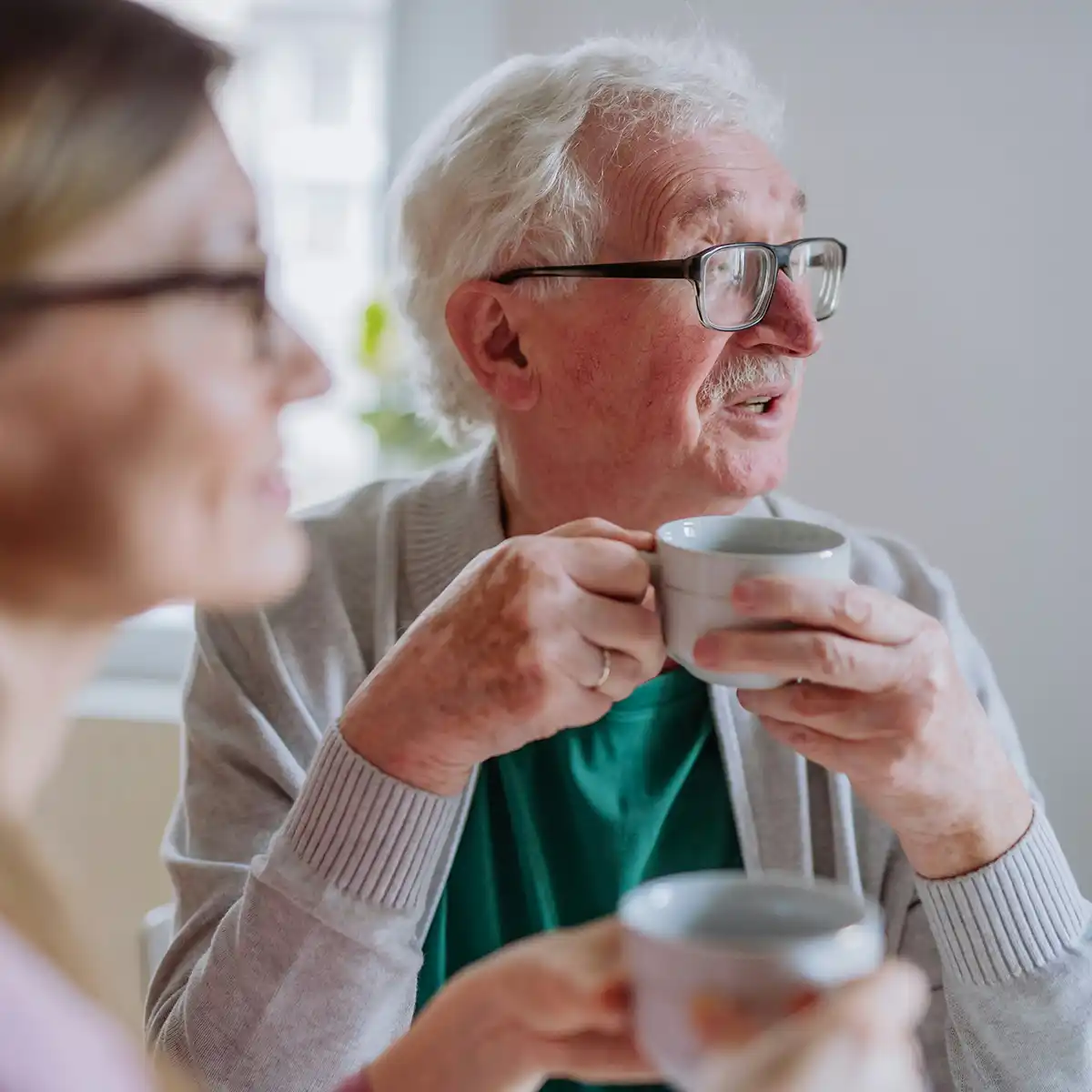 Oudere man met bril drinkt uit een kopje en zit naast een vrouw met bril die ook een kopje vasthoudt.