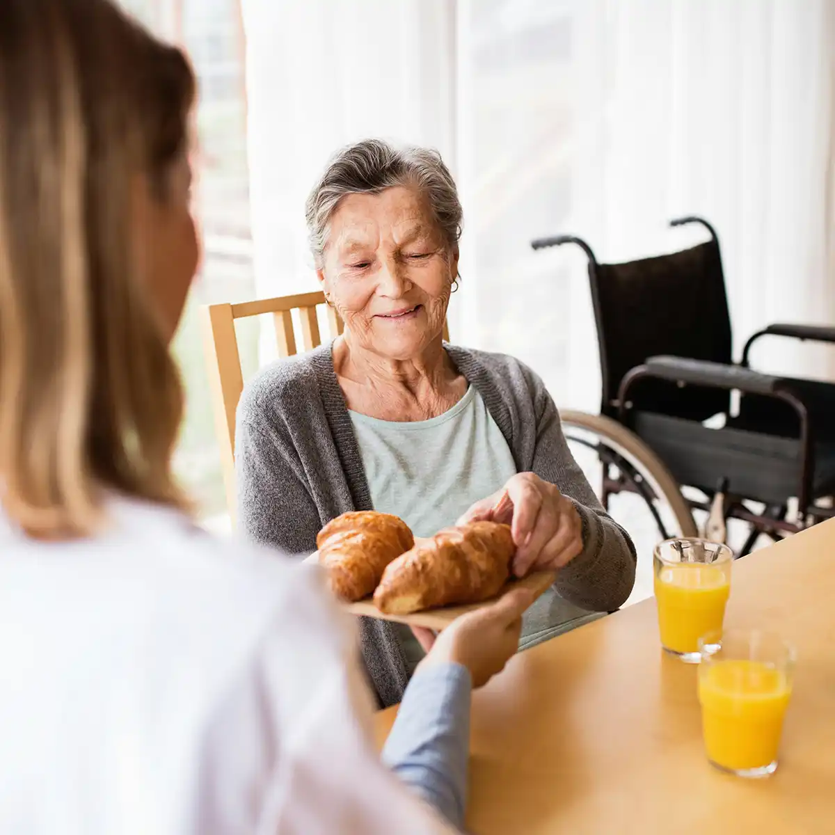 Oudere vrouw die een croissant pakt van een dienblad gehouden door een verzorger, met een rolstoel en twee glazen sinaasappelsap op tafel.