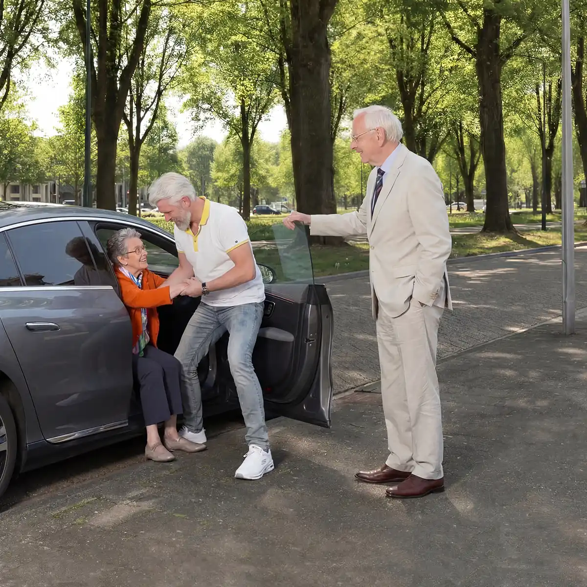 Een oudere vrouw die uit een grijze auto stapt met hulp van een man in een wit shirt, terwijl een oudere man in een beige pak de autodeur openhoudt in een zonnig park.