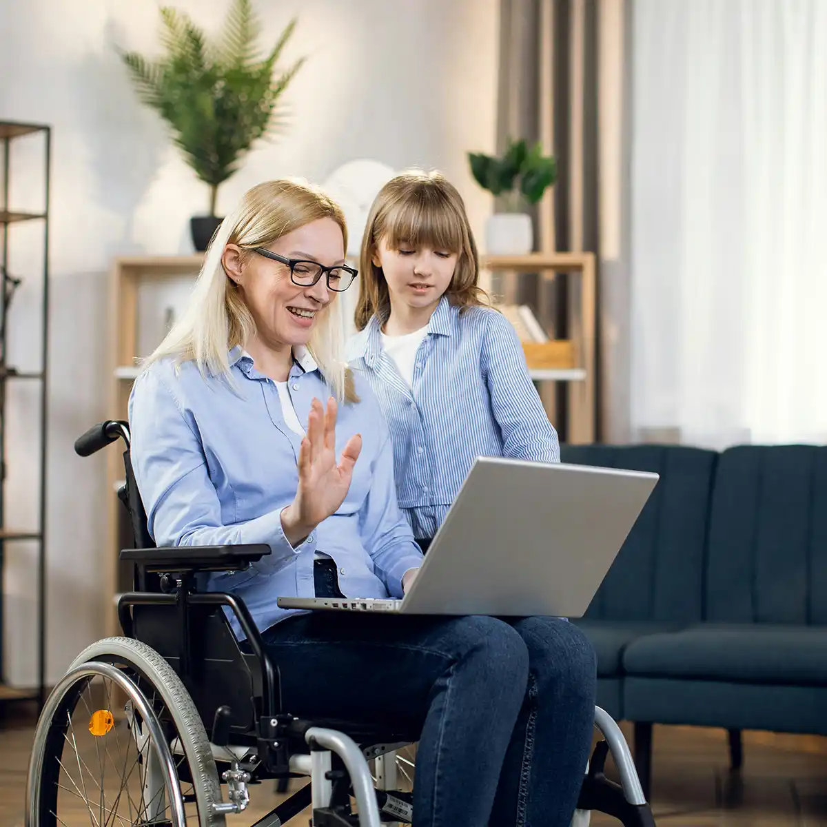 Vrouw in rolstoel zwaaiend naar een laptop terwijl een jong meisje naast haar staat in een gezellige woonkamer.