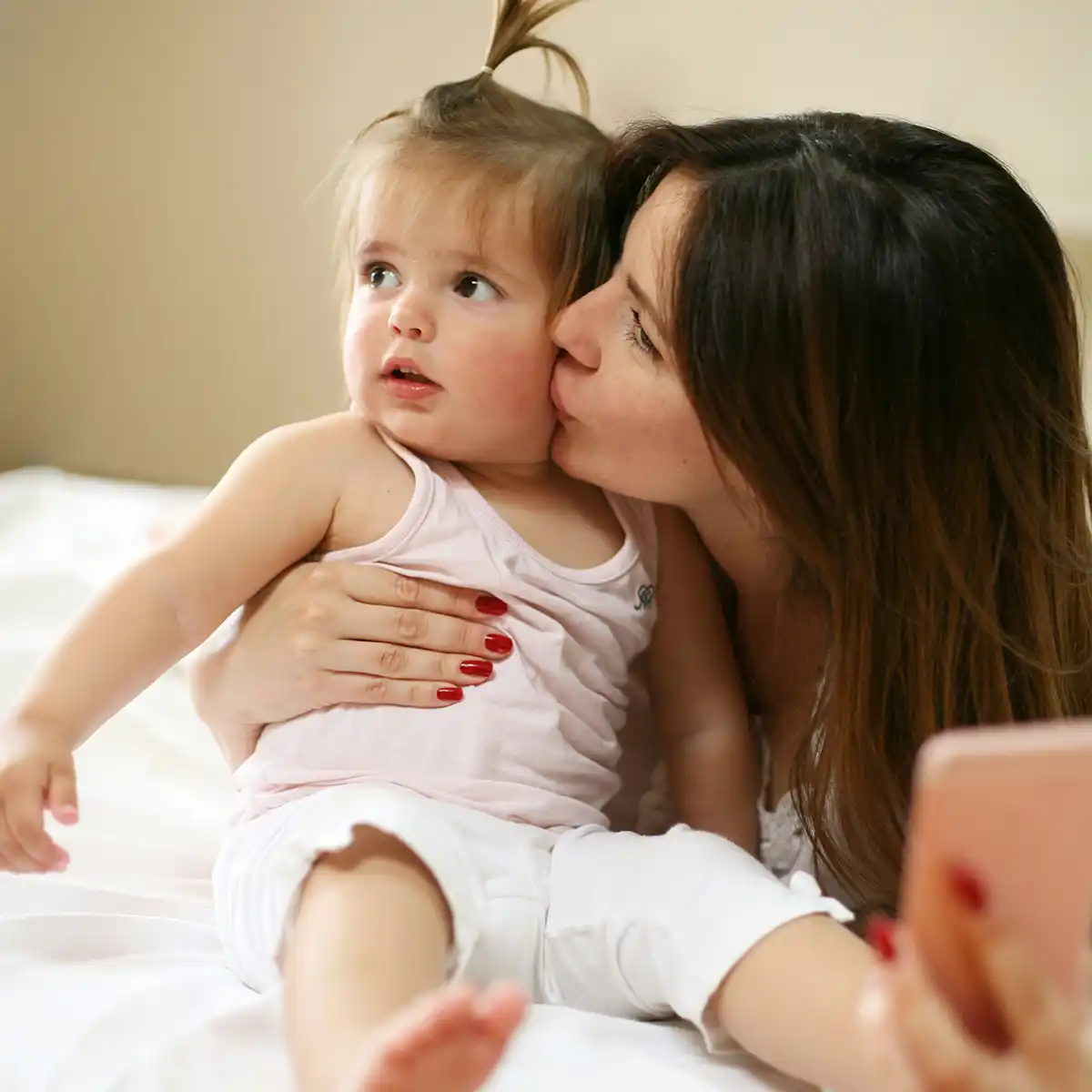 Moeder kust liefkozende haar jonge dochter die op bed zit terwijl ze een foto maakt met een smartphone.