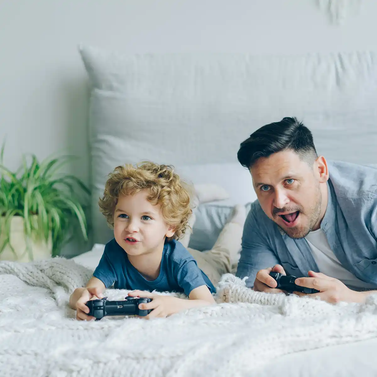 Vader en zoon liggen op bed en spelen samen een videogame met controllers in hun handen.