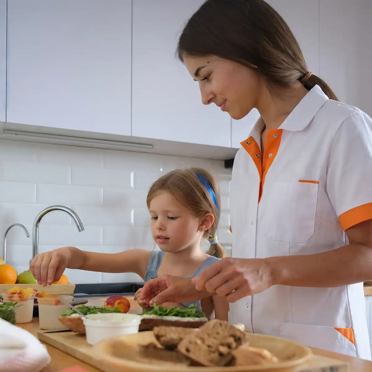Vrouw in witte en oranje uniform helpt jong meisje met het bereiden van een gezonde maaltijd in de keuken.