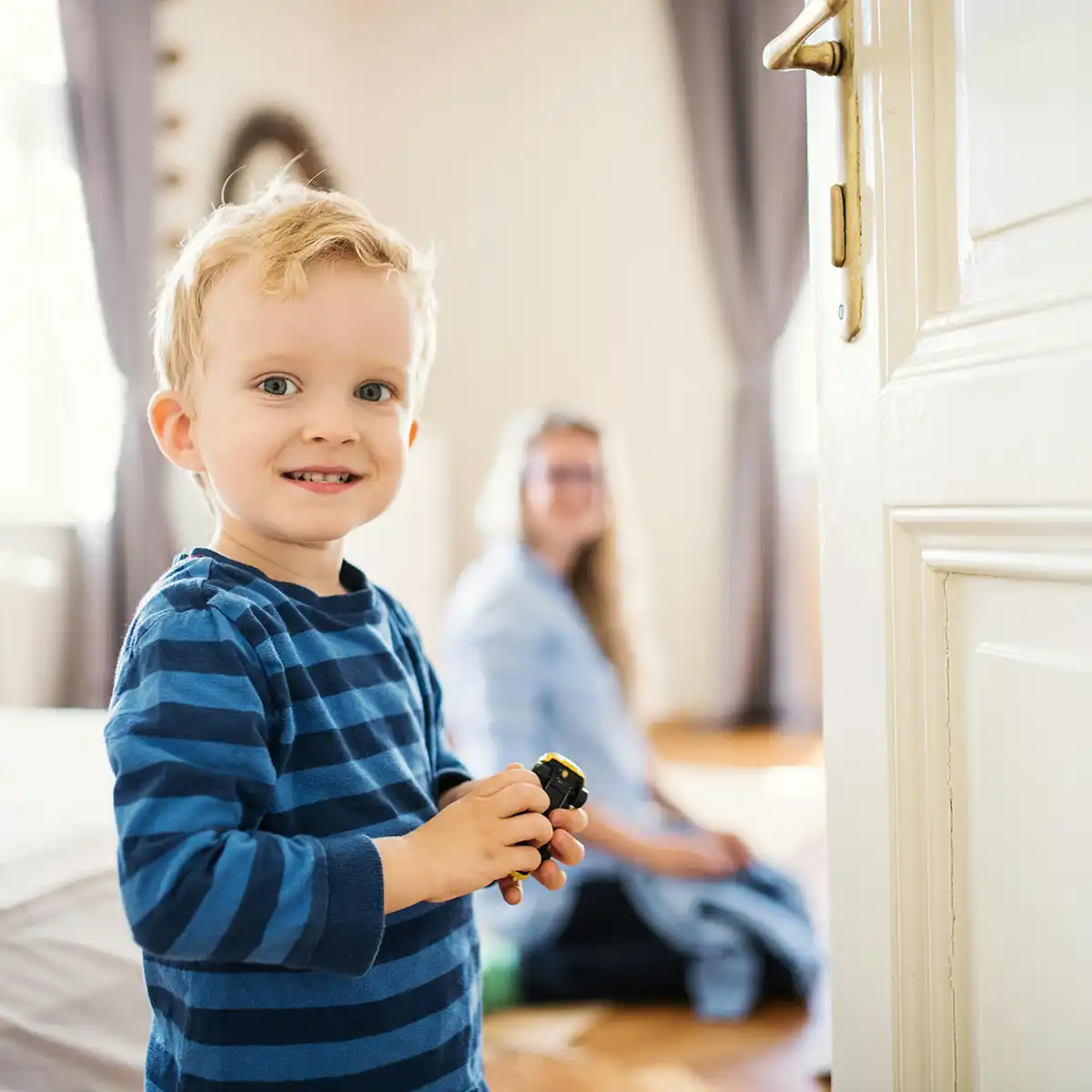 Jonge jongen met een blauw gestreept shirt die een speelgoedauto vasthoudt, met een vrouw die op de achtergrond zit.