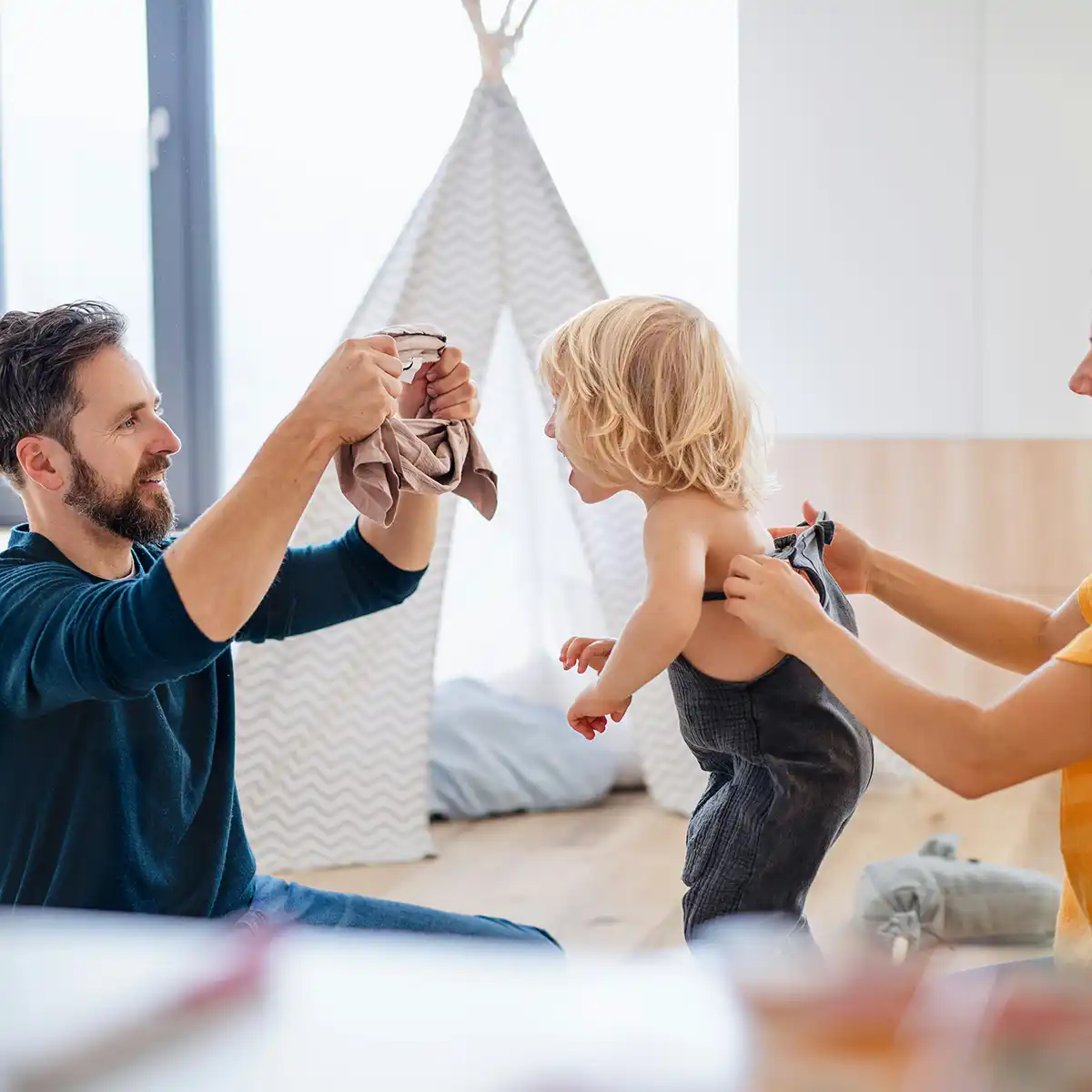 Vader en moeder helpen een peuter zich aan te kleden in een lichte woonkamer met een speeltent op de achtergrond.