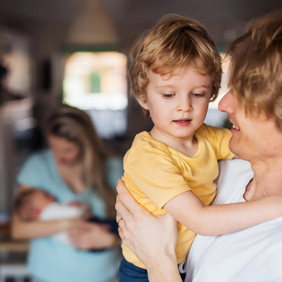 Vader houdt zijn jonge zoon in zijn armen terwijl moeder op de achtergrond hun baby vasthoudt.