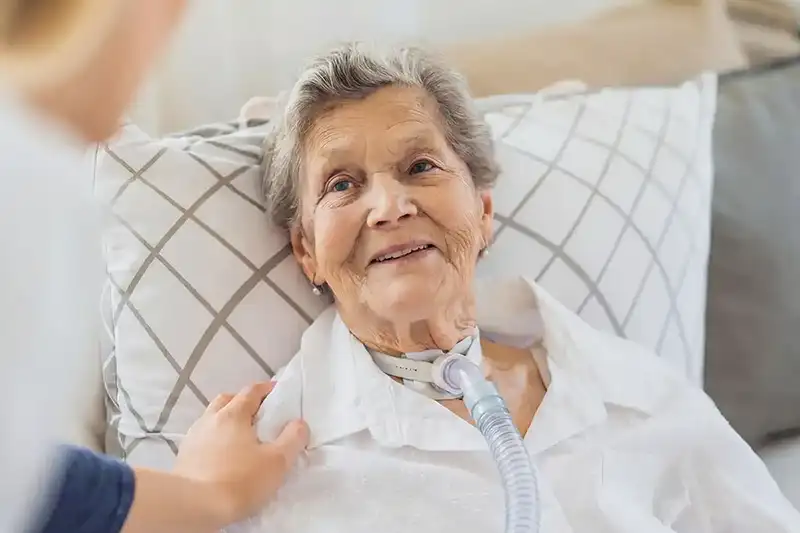 Oudere vrouw liggend op bed met tracheostomie en ademhalingsbuis, terwijl een persoon haar bemoedigend aanraakt.