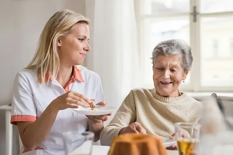 Verzorgende voert een oudere vrouw die samen aan een tafel zitten, glimlachend en genietend van eten.
