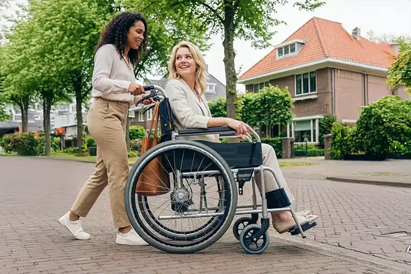 Een vrouw duwt een glimlachende vrouw in een rolstoel over een straat in een woonwijk.