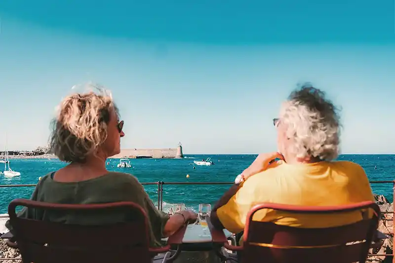 Twee oudere personen zitten buiten bij een tafel met uitzicht op een blauw meer met boten en een vuurtoren in de verte.