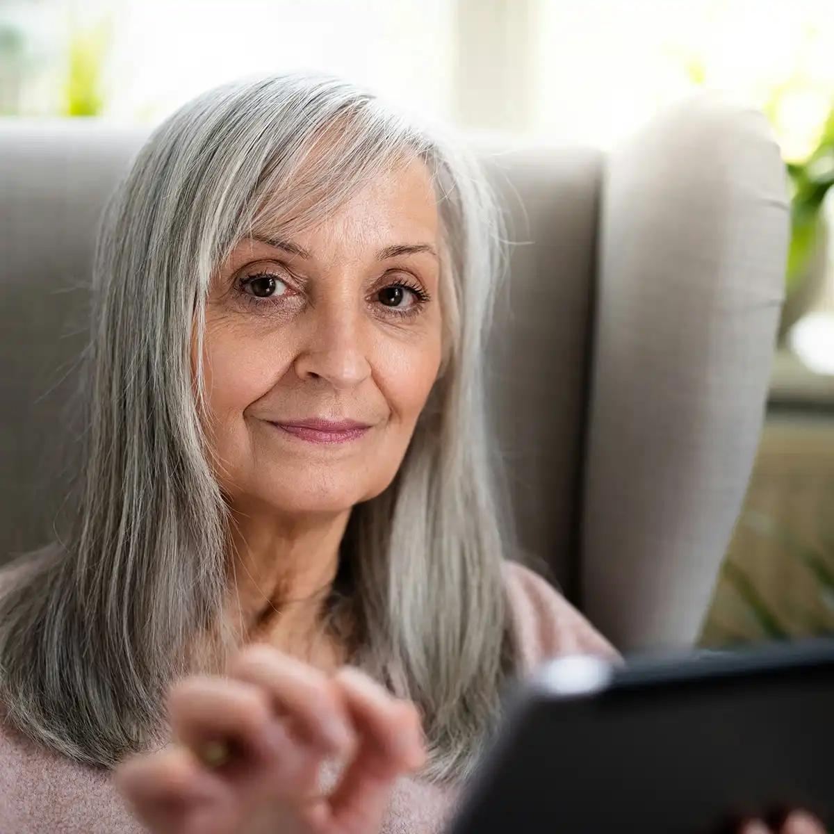 Oudere vrouw met grijs haar die een tablet gebruikt, zittend in een comfortabele stoel.