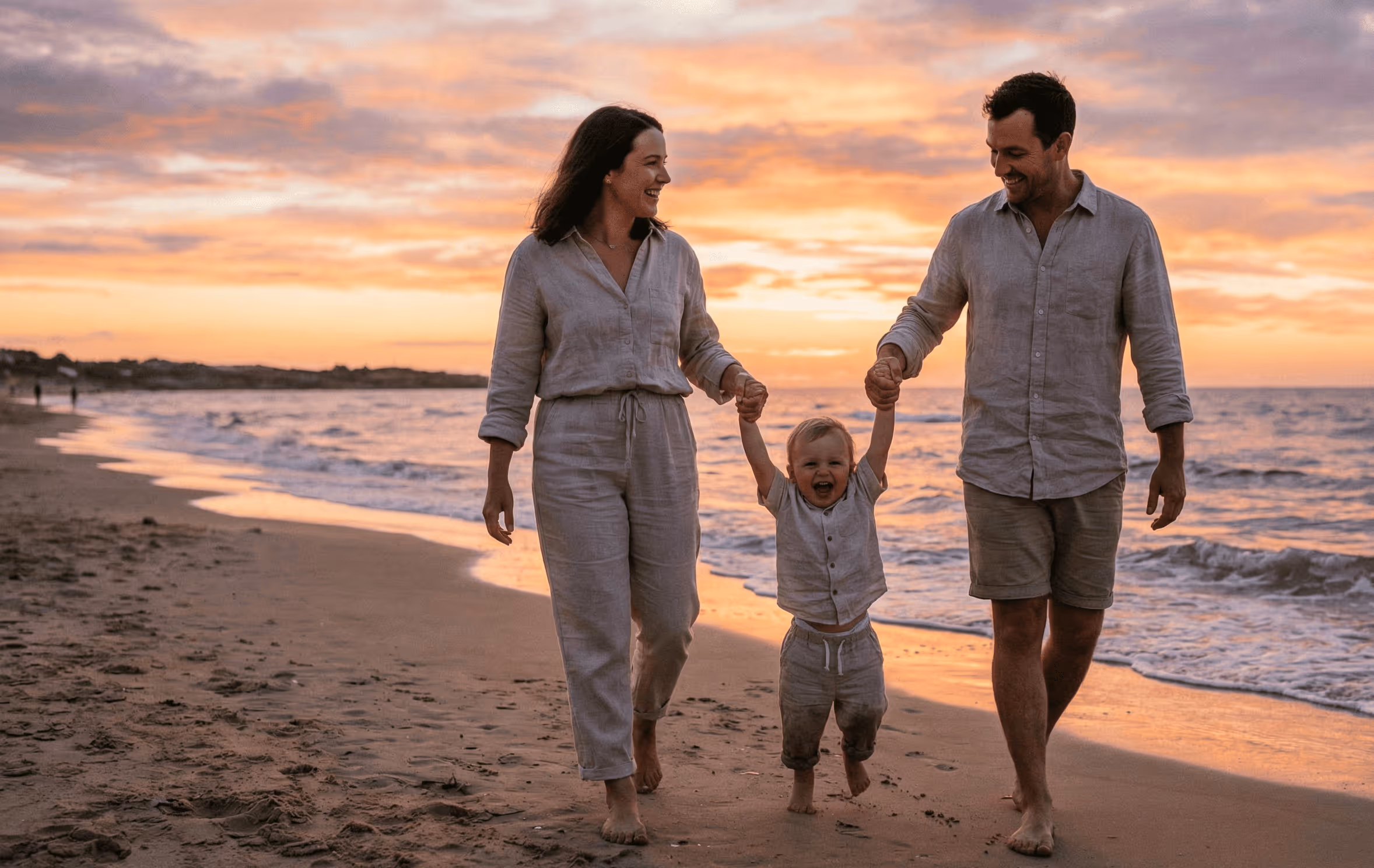 Eltern halten lachendes Kleinkind an den Händen und gehen barfuß am Strand bei Sonnenuntergang.