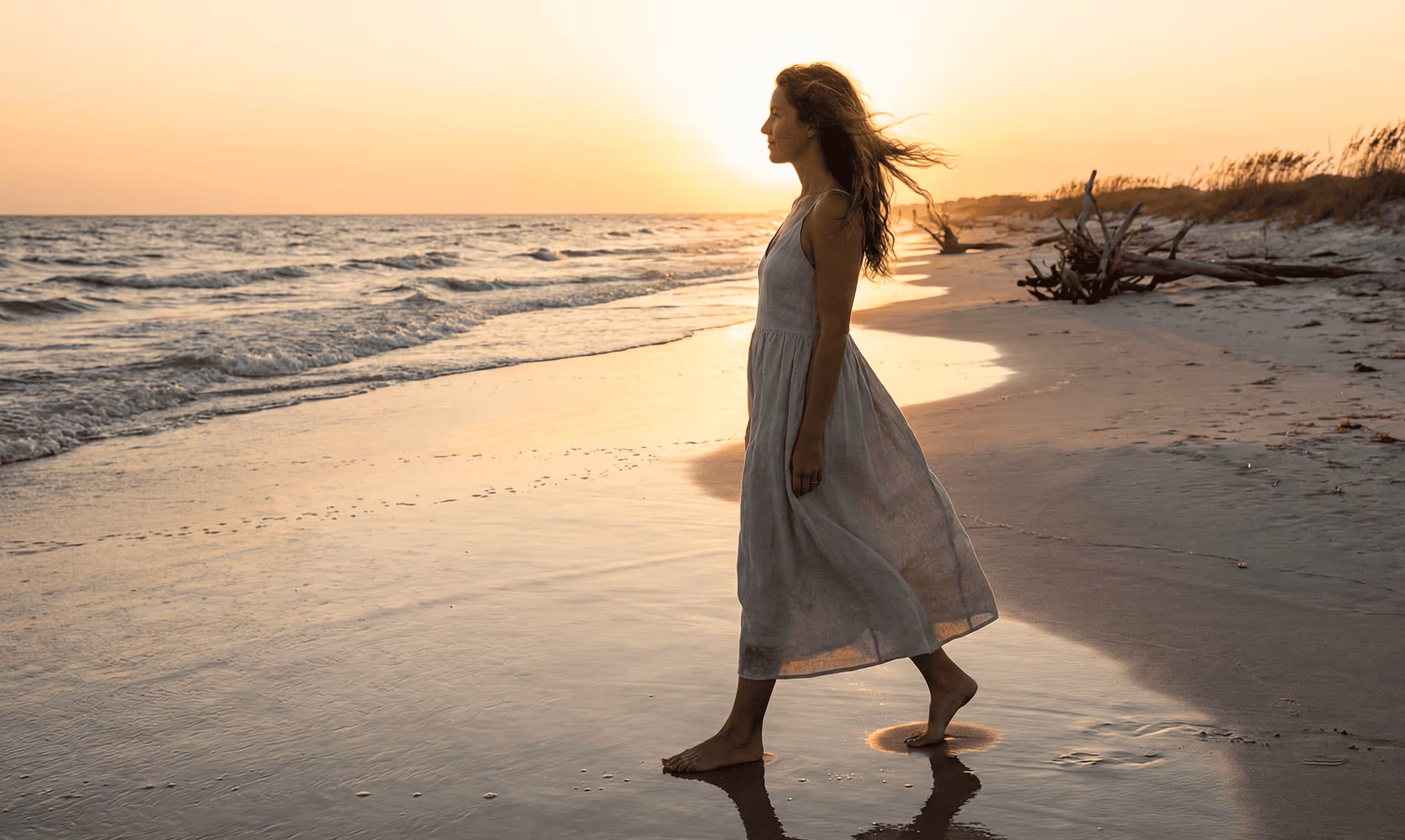 Frau in einem leichten Kleid geht barfuß am Strand entlang bei Sonnenuntergang.