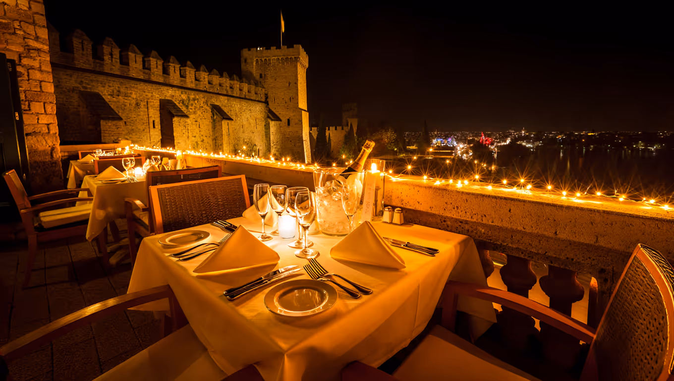 Elegant outdoor dining table set for four with white linens, wine glasses, and candles on a terrace at night overlooking a lit stone castle wall and city lights.