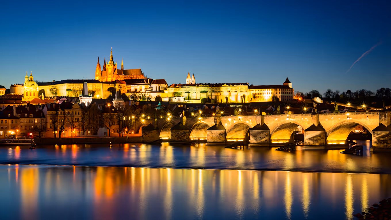 Illuminated Prague Castle and Charles Bridge at dusk reflected in the Vltava River.