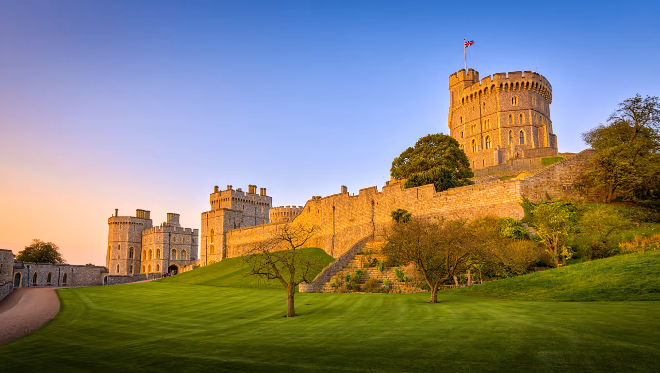Windsor Castle illuminated by golden sunlight with green lawns and sparse trees under a clear blue sky at sunset.