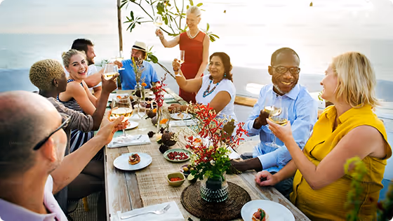 Diverse group of people enjoying an outdoor meal with drinks and food, smiling and toasting at a wooden table near water.