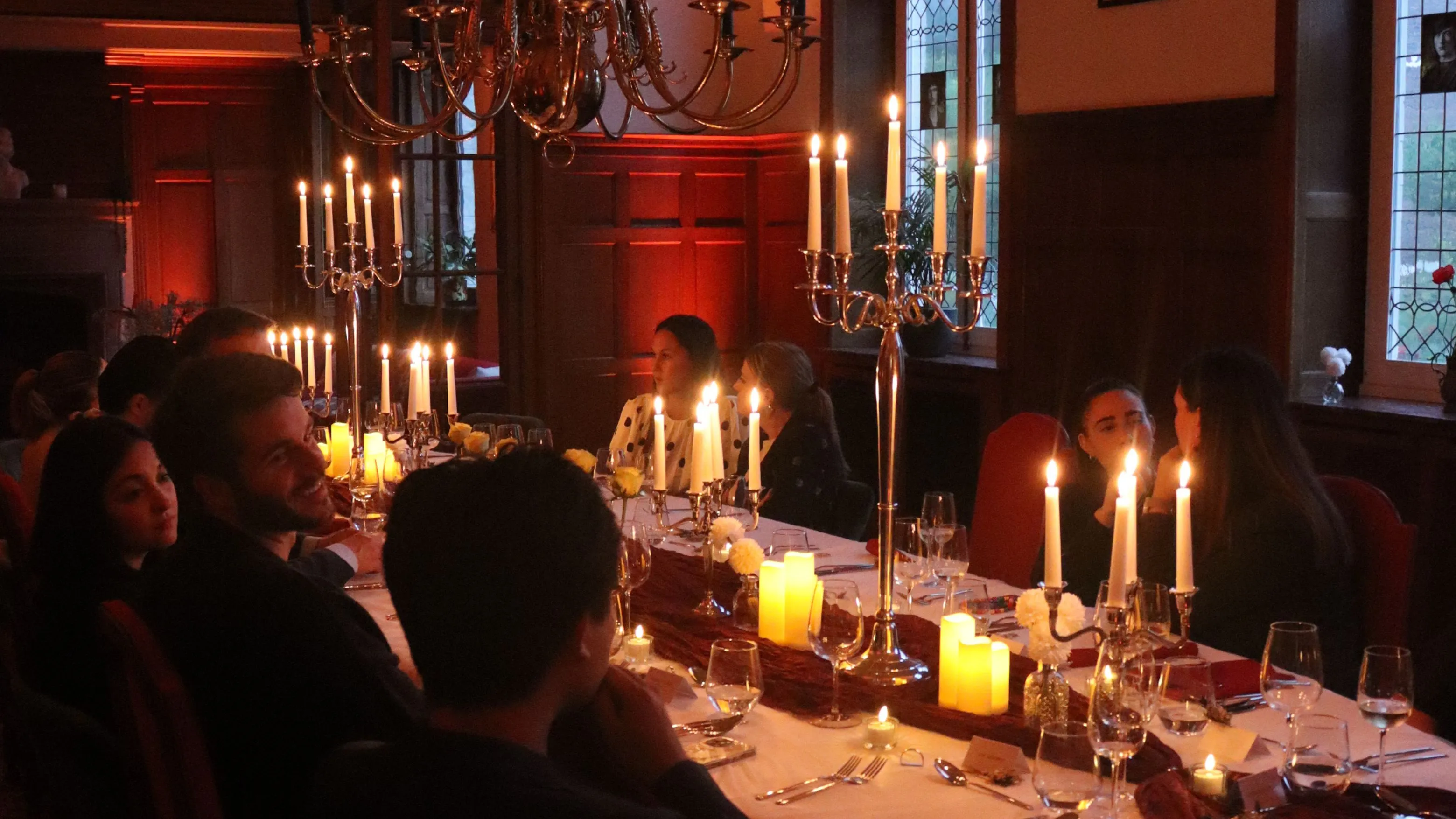 Long, elegantly set banquet table with lit candles in a grand hall featuring stained glass windows and banners on the walls.