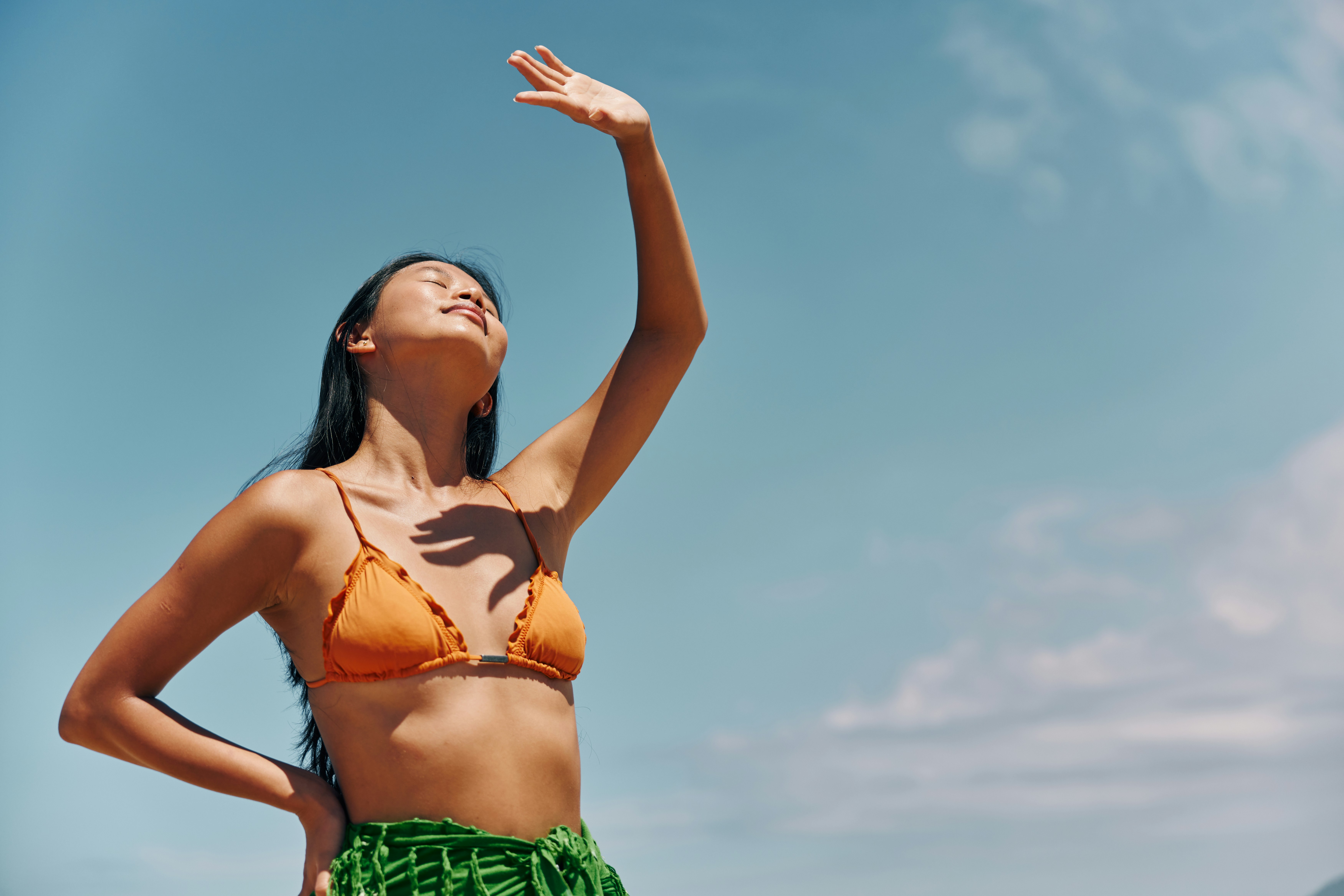 Frau im orangefarbenen Bikinioberteil hebt einen Arm in die Sonne vor blauem Himmel.